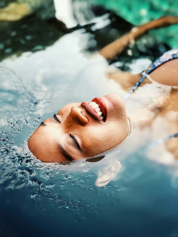 A young woman half-submerged in a pool of water, face-up, relaxing with eyes closed